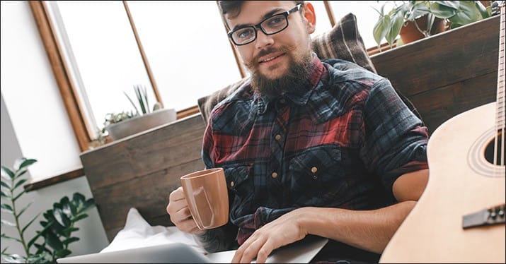Relaxed man with glasses and a beard enjoying coffee and using a laptop in a cozy home office setting, with an acoustic guitar on the side, surrounded by houseplants and natural light.