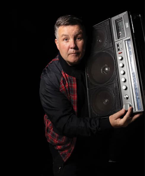 Vintage boombox speaker held by a man with short dark hair, wearing a black jacket with red plaid accents, against a black background.
