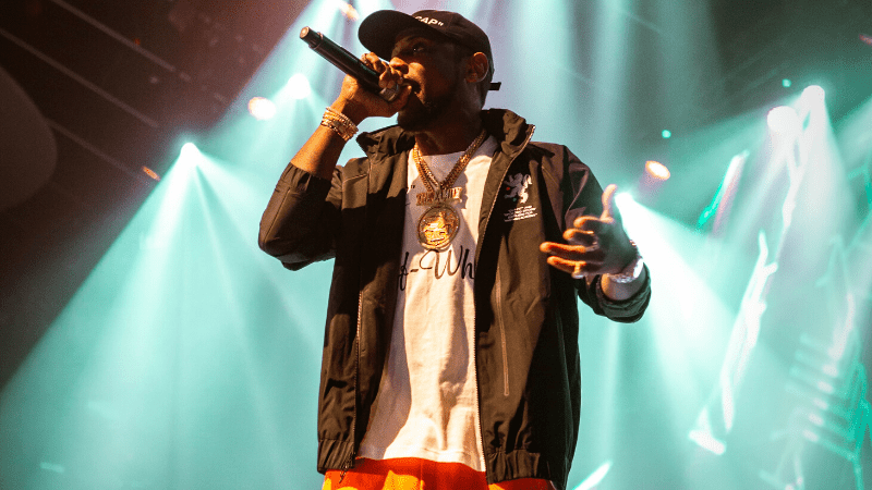 Rapper performing on stage with microphone, vibrant concert lighting, hip hop artist, live music event, urban music performance, Hypebot music industry news.