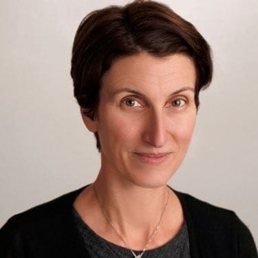 A professional woman with short dark hair and a friendly expression, wearing a black blazer and a necklace, against a neutral background.