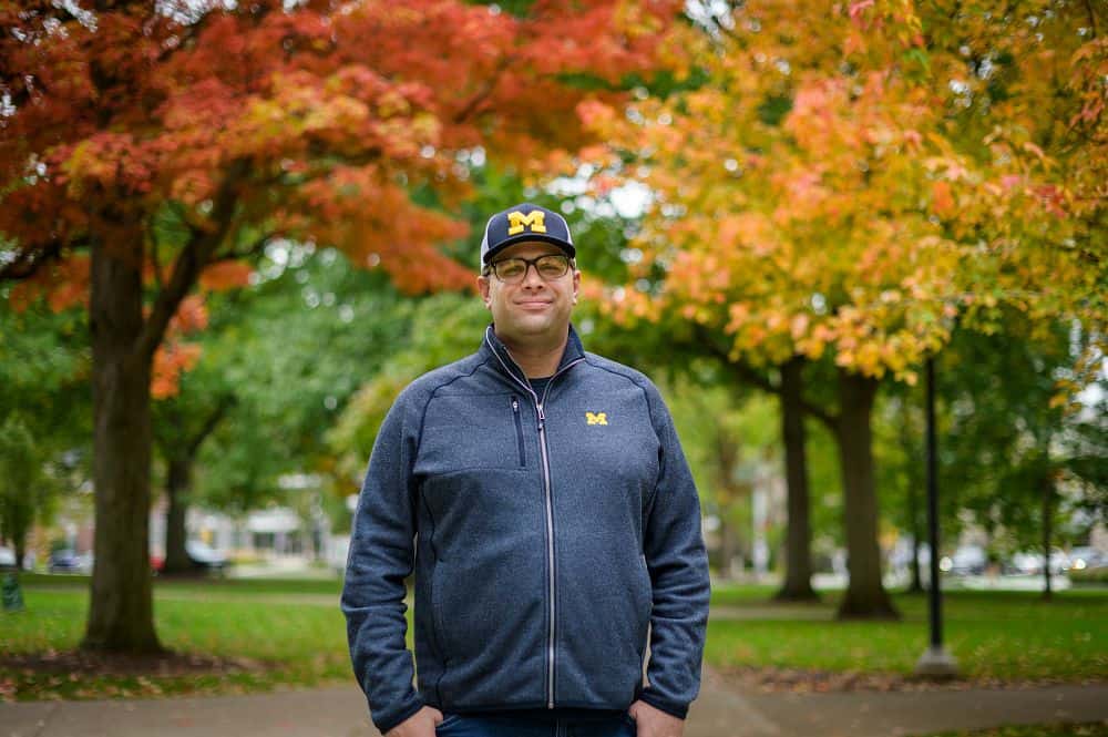 Michigan Wolverines fan wearing team apparel outdoors during autumn with colorful fall foliage in the background.