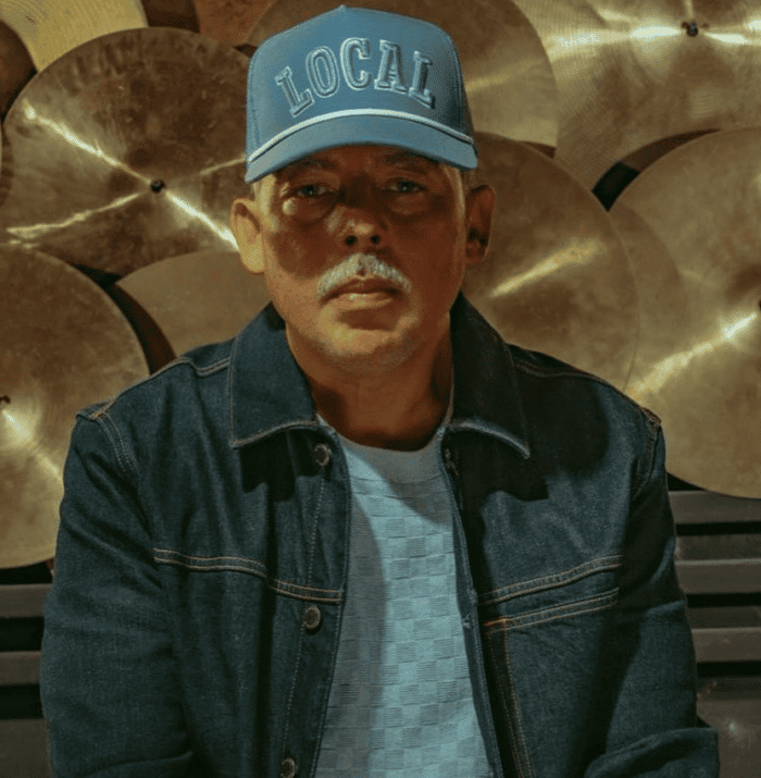 Local man wearing denim jacket and baseball cap with "LOCAL" embroidered, standing in front of a wall of cymbals, musician in a music studio, casual style, music environment, confident demeanor, streetwear fashion, professional musician, musical instruments, urban vibe.