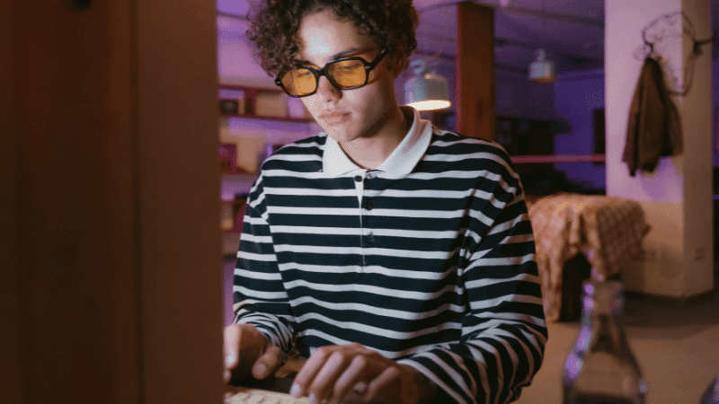 Young person wearing glasses and striped shirt working on a tablet in a cozy, dimly lit modern workspace with creative vibe.