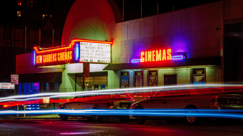 New Gardens Cinemas neon signs illuminated at night with colorful light streaks from passing cars, promoting a vibrant urban entertainment scene.