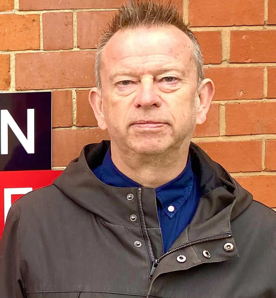 Photograph of a middle-aged man standing outdoors against a brick wall, wearing a dark jacket and blue shirt, with a serious expression, emphasizing the importance of music industry insights on Hypebot.
