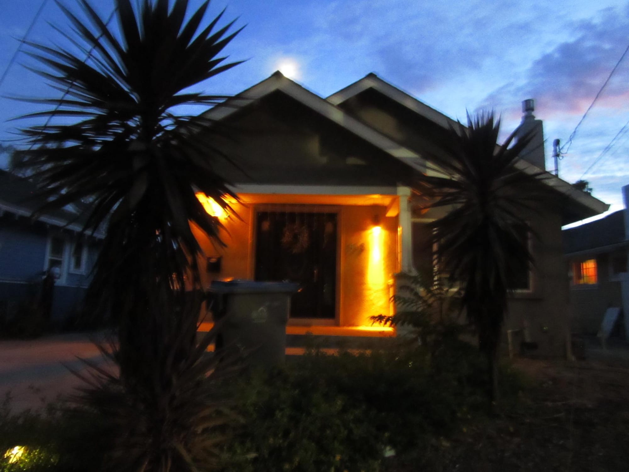 a picture of a house flooded with bright orange light against the backdrop of a dramatic, ghostly blue cloudy sky with streak of pink as the sun sets. it cuts an eerie and otherworldly atmosphore.