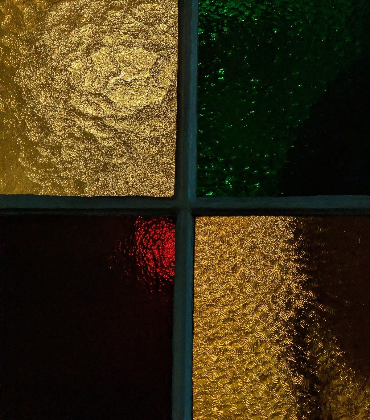 A glass windowpane divided into four quadrants; glazed in green, an opaque gold foil,  a deep blood red and a shimmering yellow 