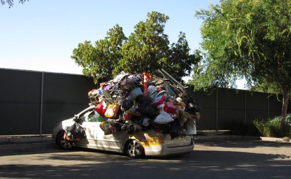 a white car in a parking lot filled to overflowing with plastic bags of different colors. The explosion of bags is in visual harmony with the tree behind the car.