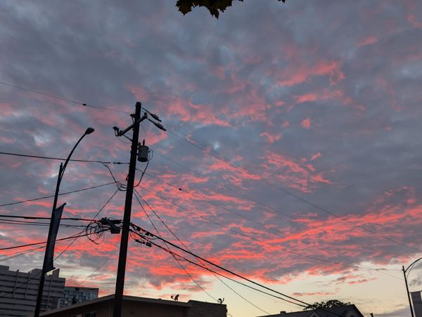 a picture of a sunset with vivid orange, red and grey clouds. From the camera angle, the powerline with wires stretched out of both sides looks tall enough to pierce the clouds
