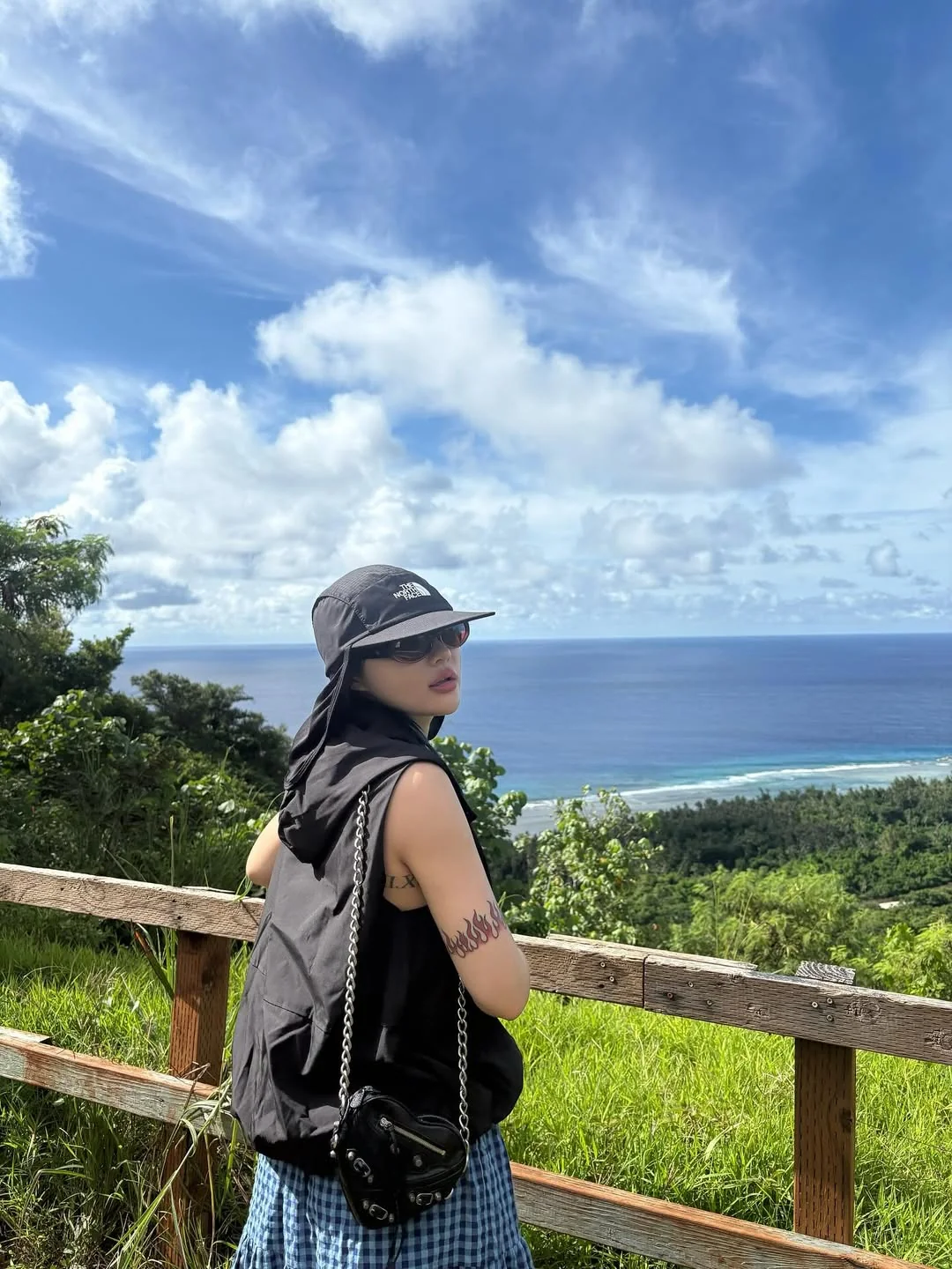 Siyeon, dressed in a black hat and sleeveless top and checkered skirt, looks out over a view of the ocean.