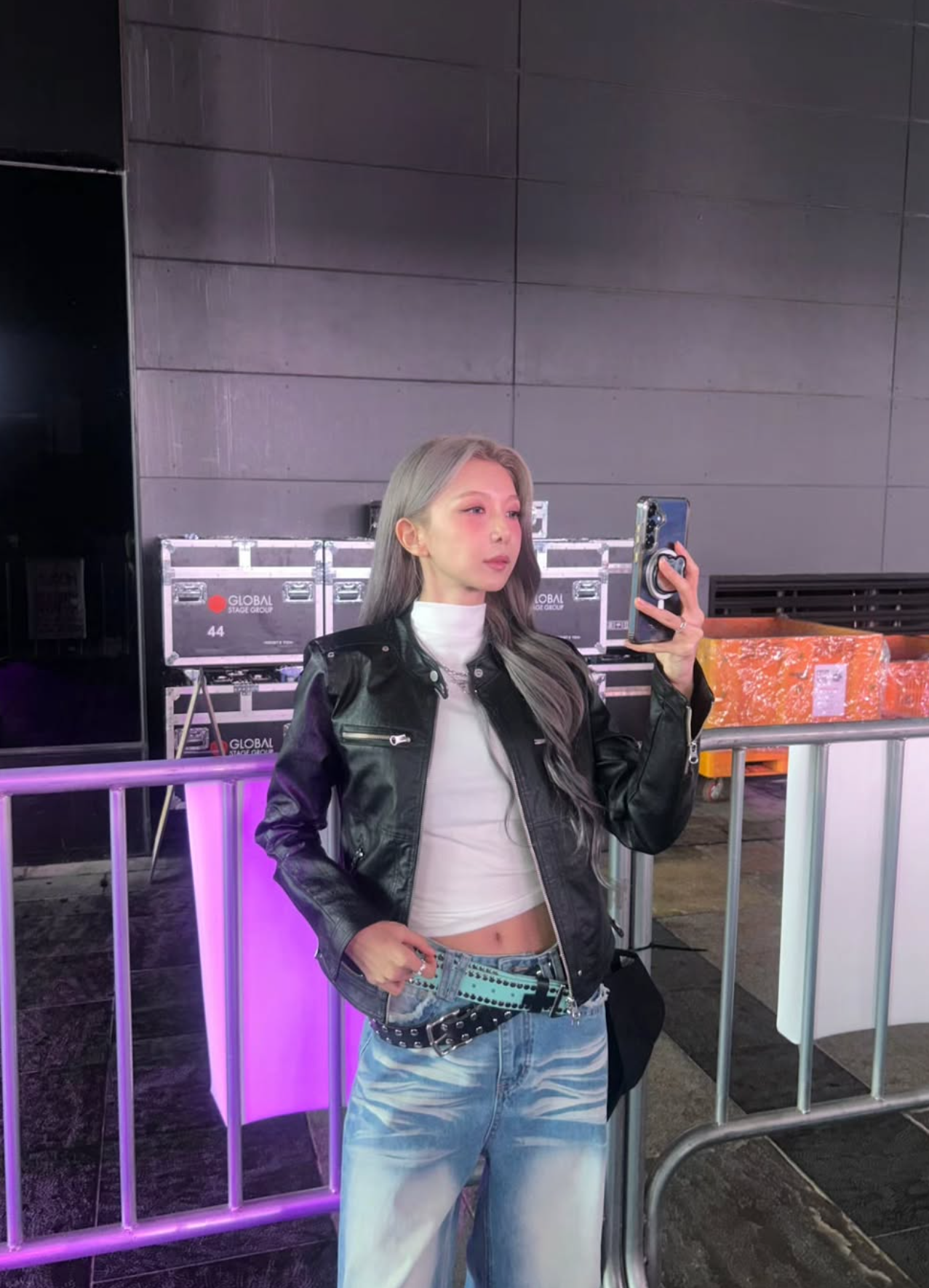 Dami in a black leather jacket, white turtleneck, and jeans taking a photo.