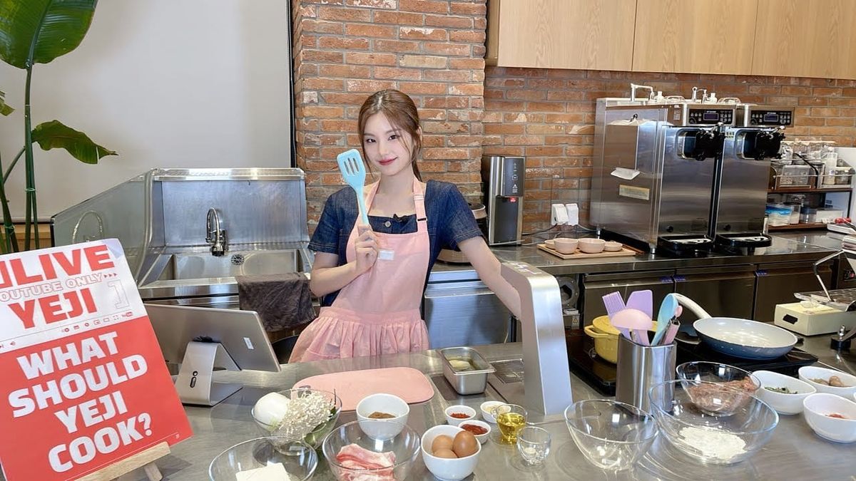 ITZY's Yeji in a pink apron smiling for the camera holding a spatula surrounded by baking ingredients