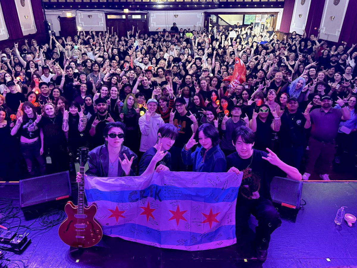 ChRocktikal posing with a Chicago flag on stage as the audience in the background cheers.