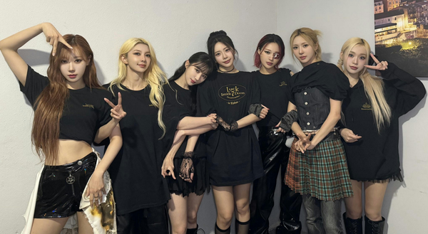 Dreamcatcher dressed in black, poses for a group picture against a white wall, smiling.