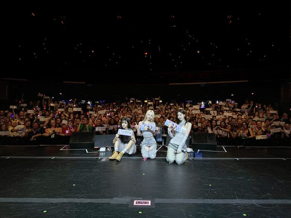 Dreamcatcher UAU's JiU, SuA, and Yoohyeon take a picture with fans in the background holding banners.