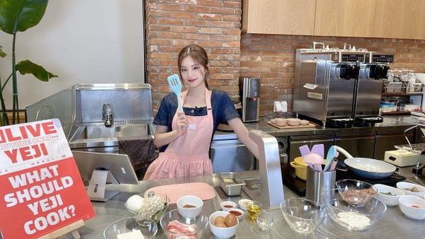 ITZY's Yeji in a pink apron smiling for the camera holding a spatula surrounded by baking ingredients