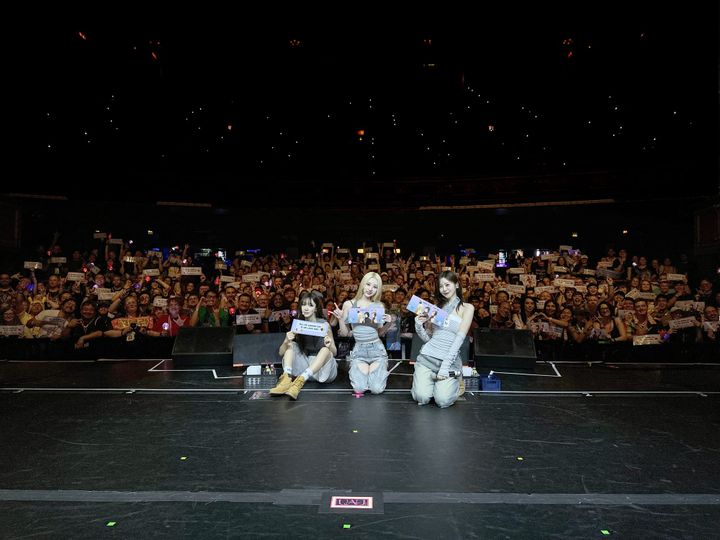 Dreamcatcher UAU's JiU, SuA, and Yoohyeon take a picture with fans in the background holding banners.
