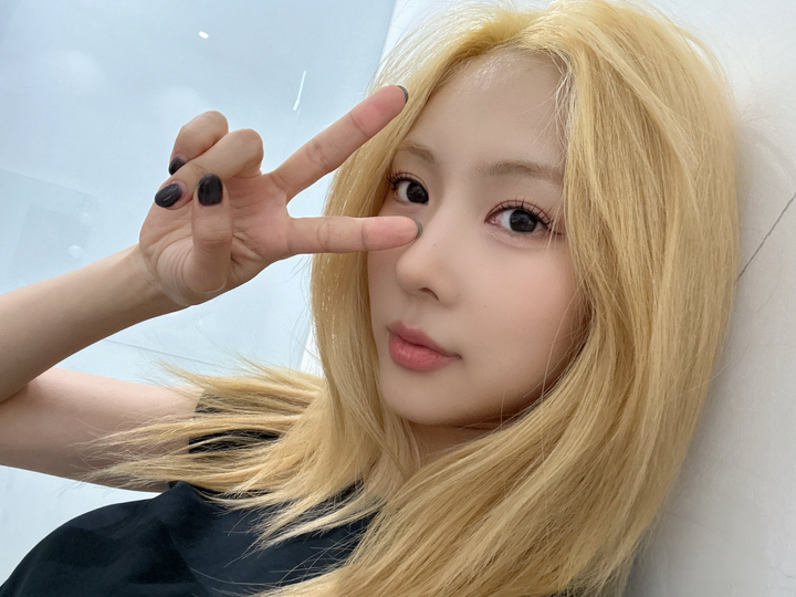 Yoohyeon with blonde hair and a black t-shirt with a V-sign towards the camera.