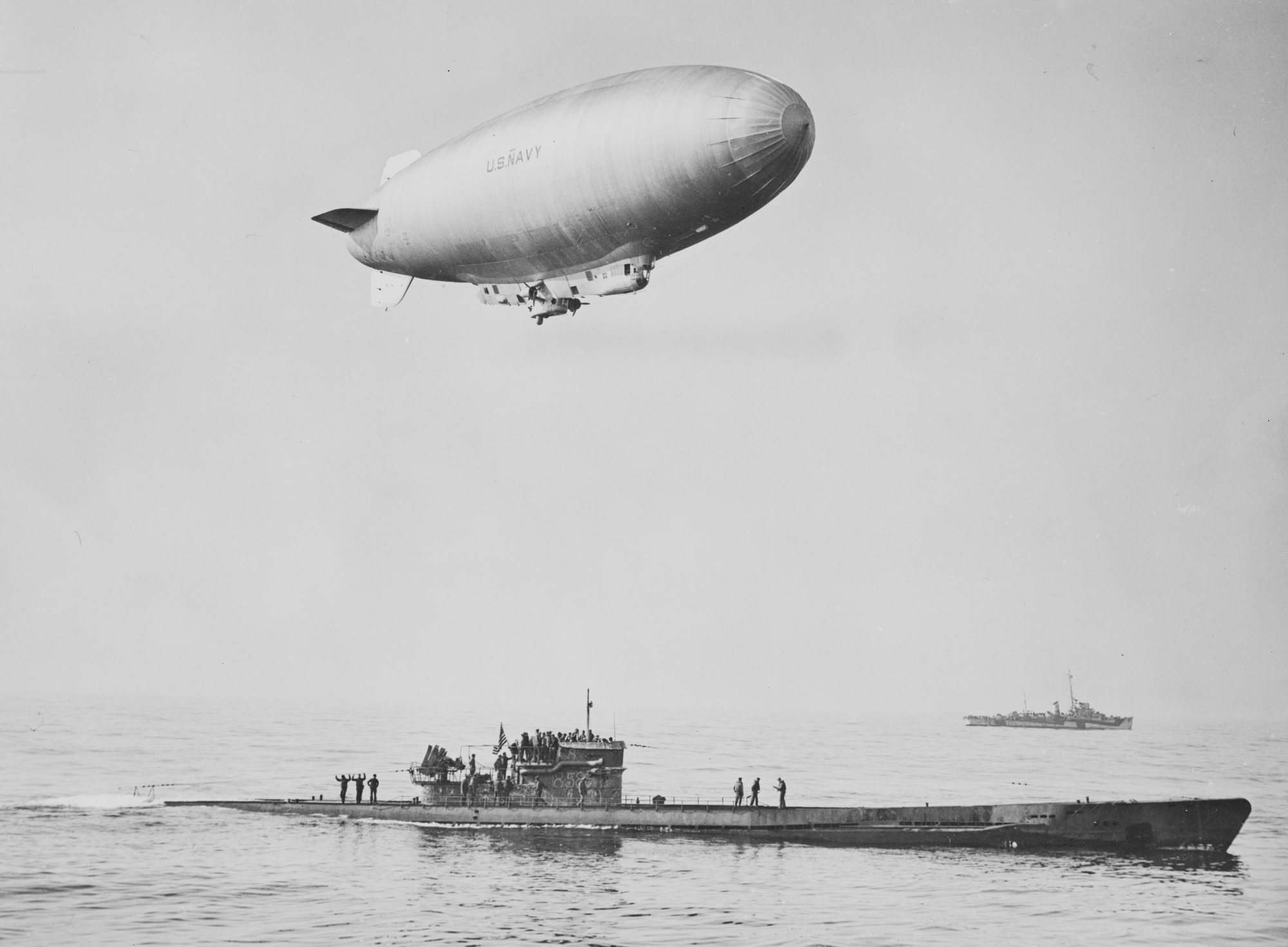 A black and white photo of a blimp over a Navy ship