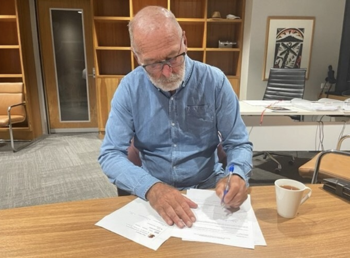 Wayne brown in a blue shirt with a cup of tea next to him as he signs