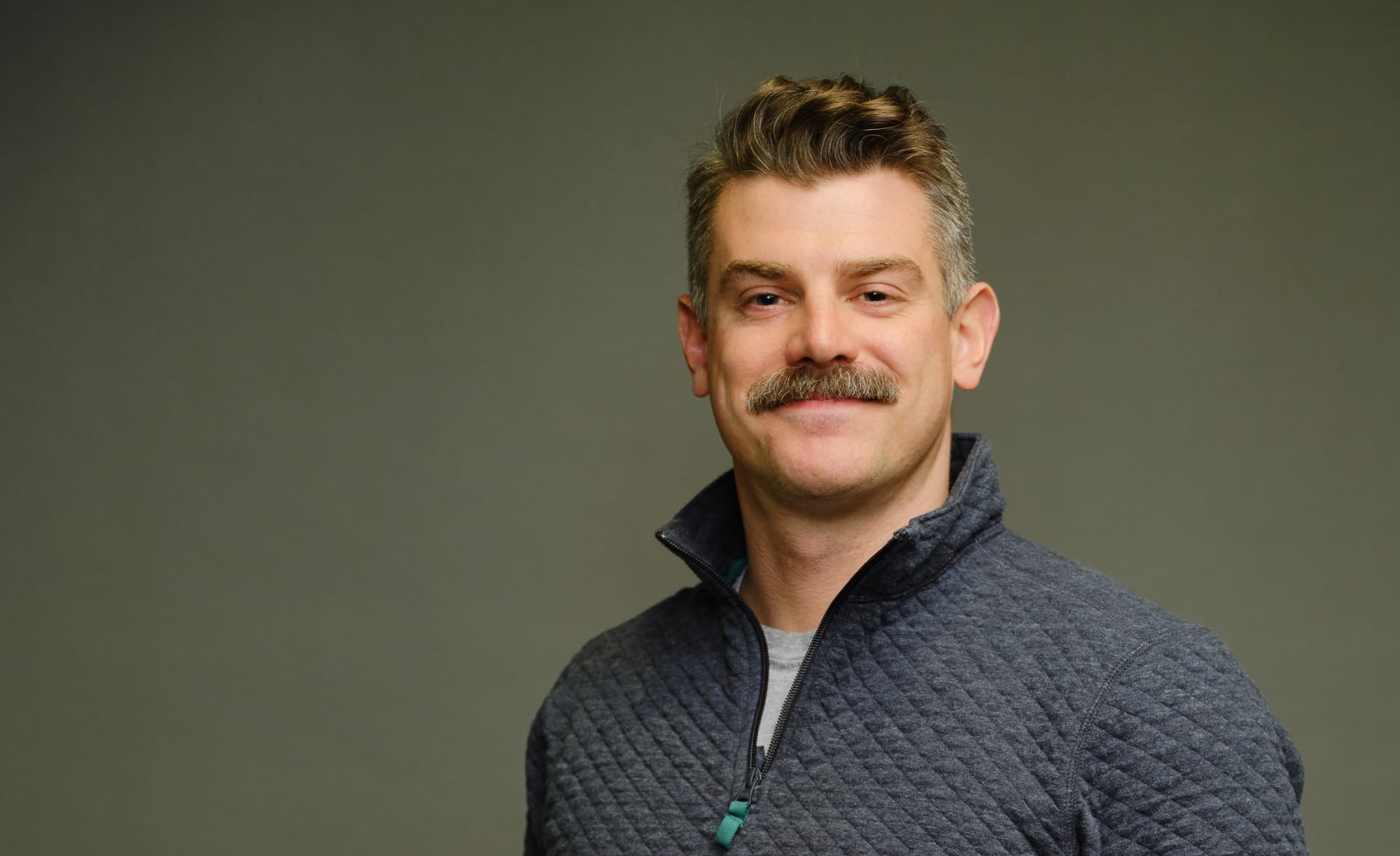 A portrait of film scholar PT Klein wearing a blue sweater and grey shirt against an olive background.