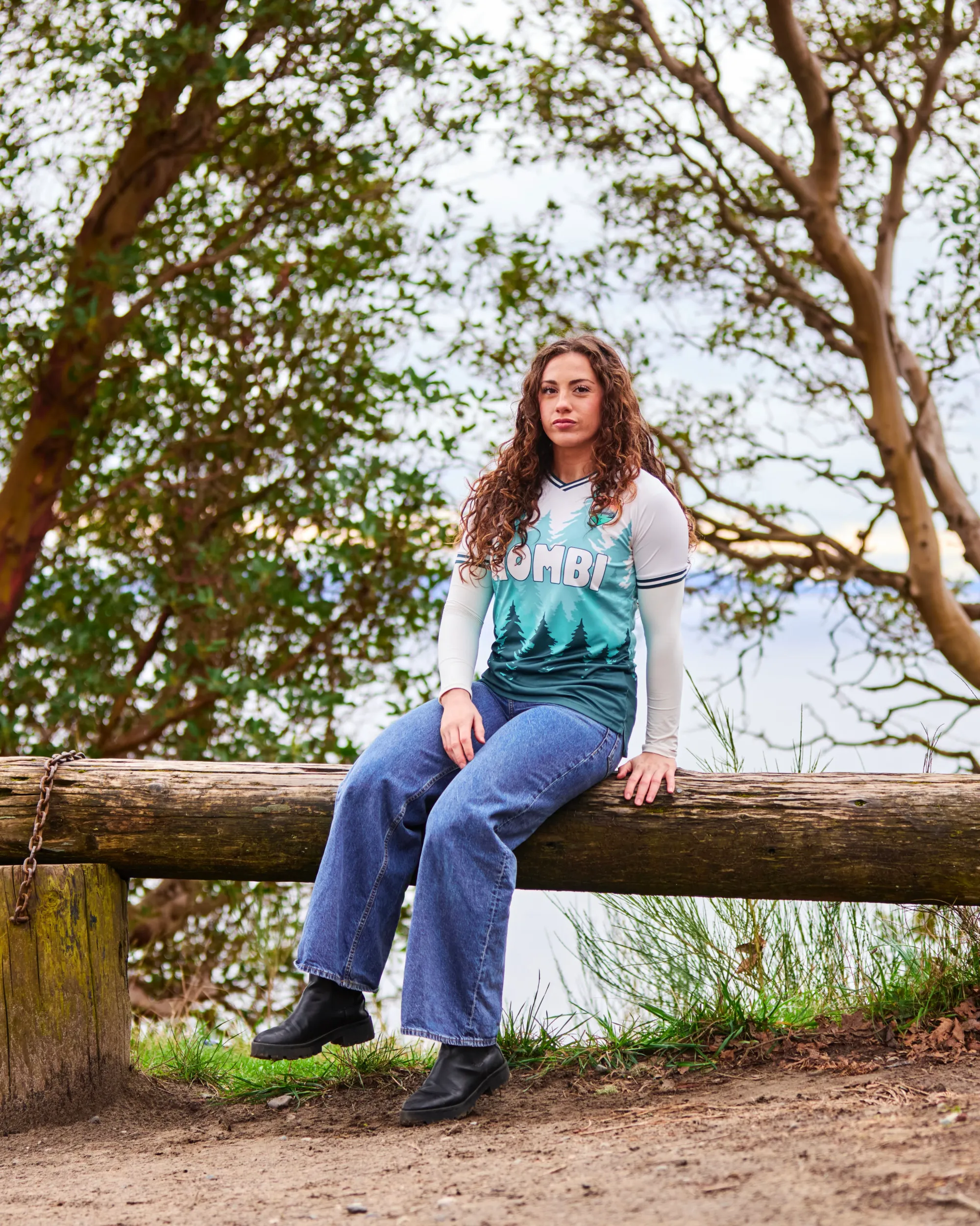 A woman sits on a log wearing a jersey with trees on it.