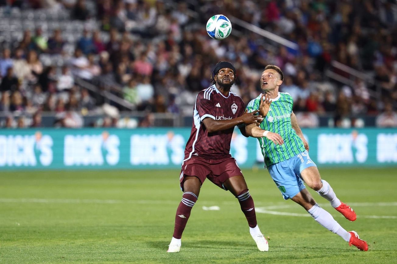 Danny Musovski in a green and blue Sounders kit challenges Chidozie Awaziem in a burgundy Rapids kit for the ball.