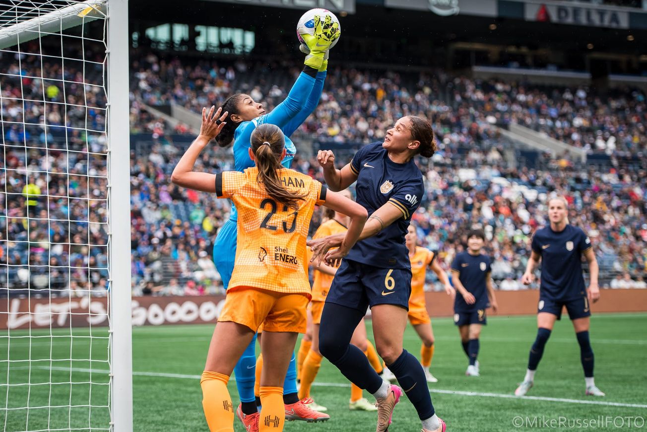 Houston Dash goalkeeper Abby Smith leaps to claim a ball over Reign forward Lynn Biyendolo and several Dash teammates in a May 2025 match.