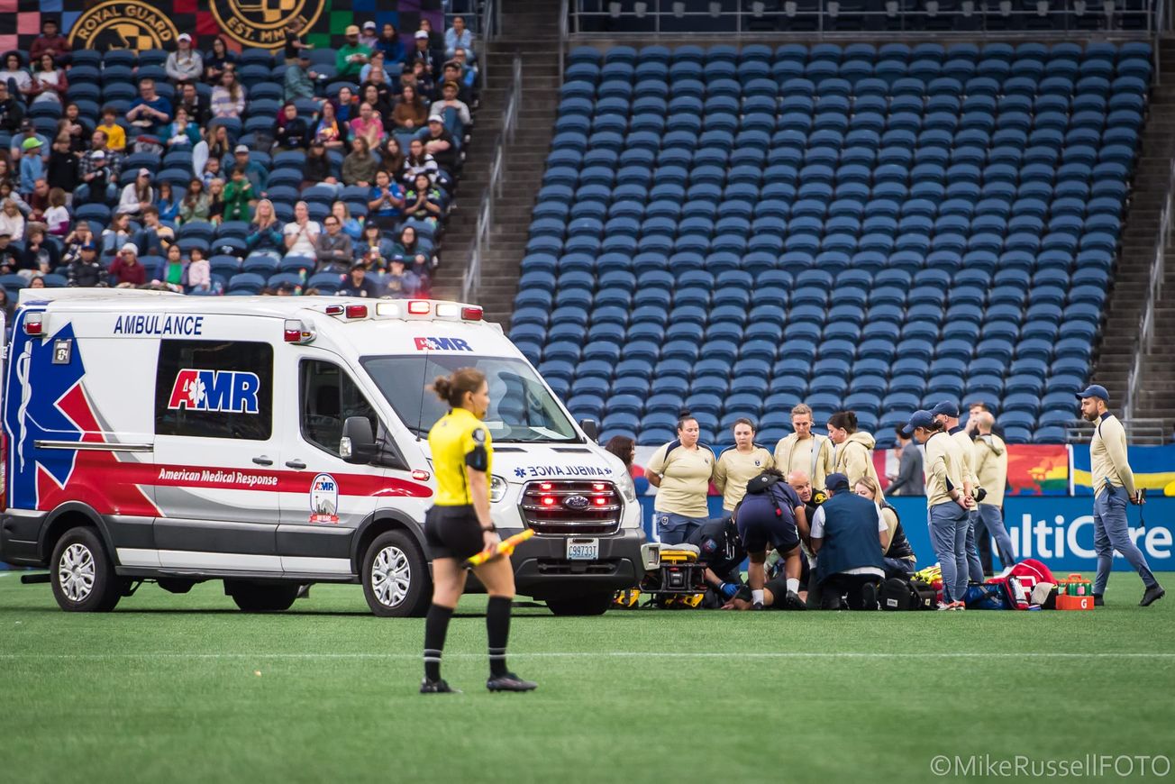 Medical personnel and team staff surround an injured player while an ambulance waits nearby during a Seattle Reign FC vs Racing Louisville match in September 2025.