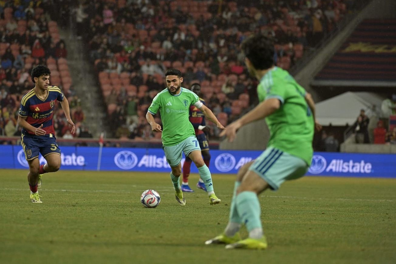 Cristian Roldan wearing a green Sounders jersey and light blue shorts dribbles with an RSL player next to him and Paul Rothrock in the foreground.