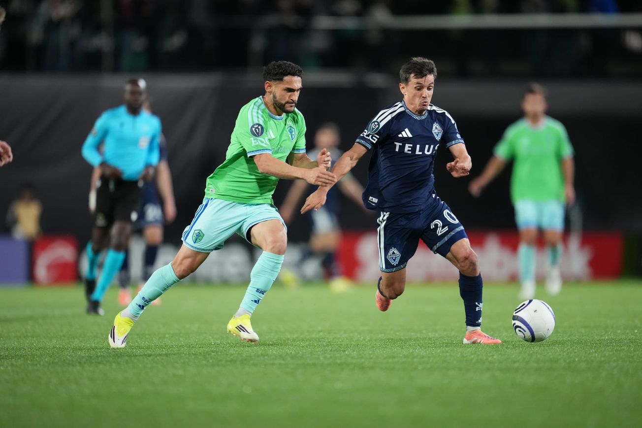 Cristian Roldan in a green Sounders jersey and light blue shorts and socks challenges a player in a dark blue Vancouver Whitecaps kit for the ball.