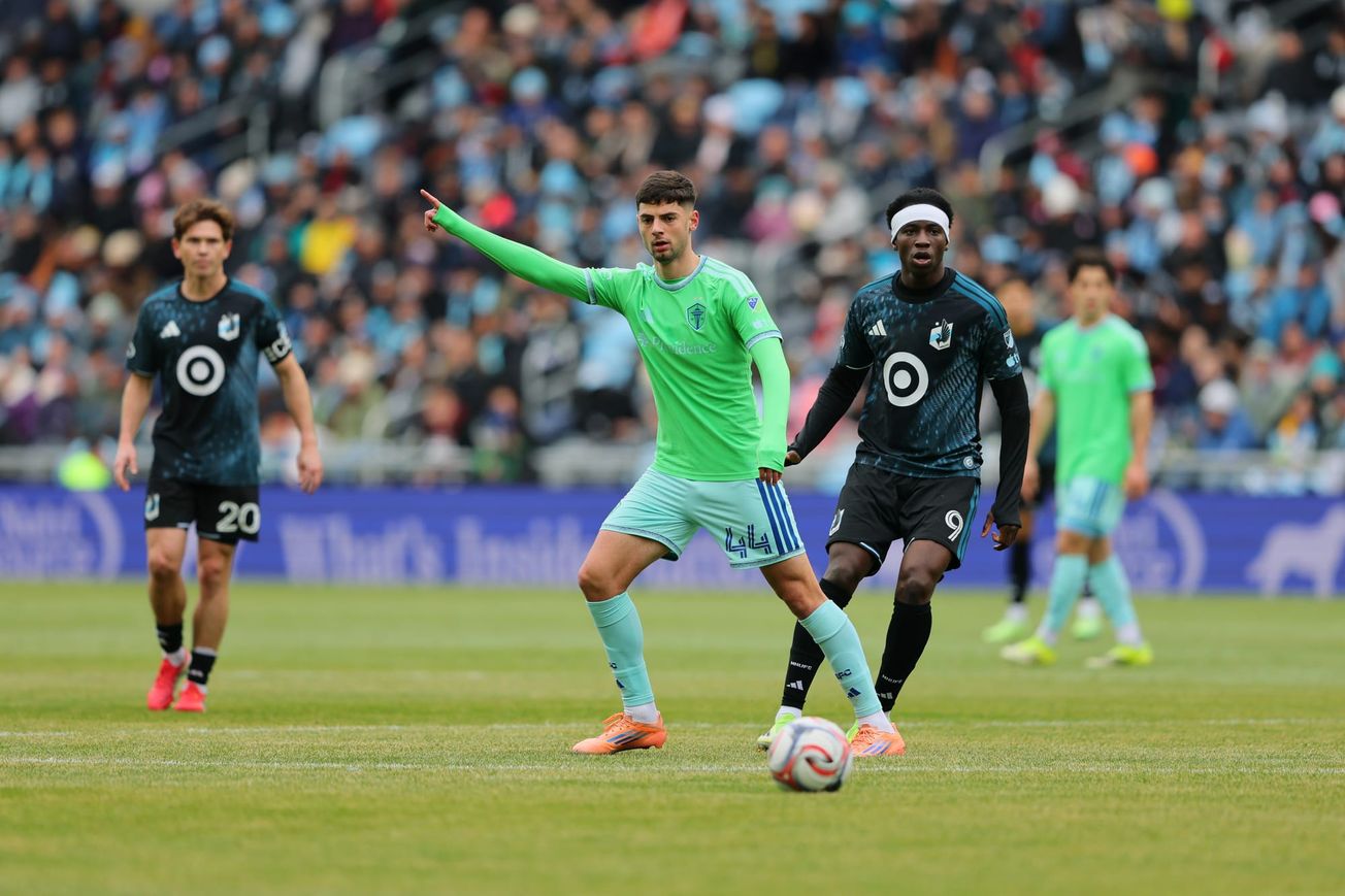 Nikola Petkovic wearing a green Sounders jersey and light blue shorts and socks with a pair of Minnesota United players in black and blue behind him.