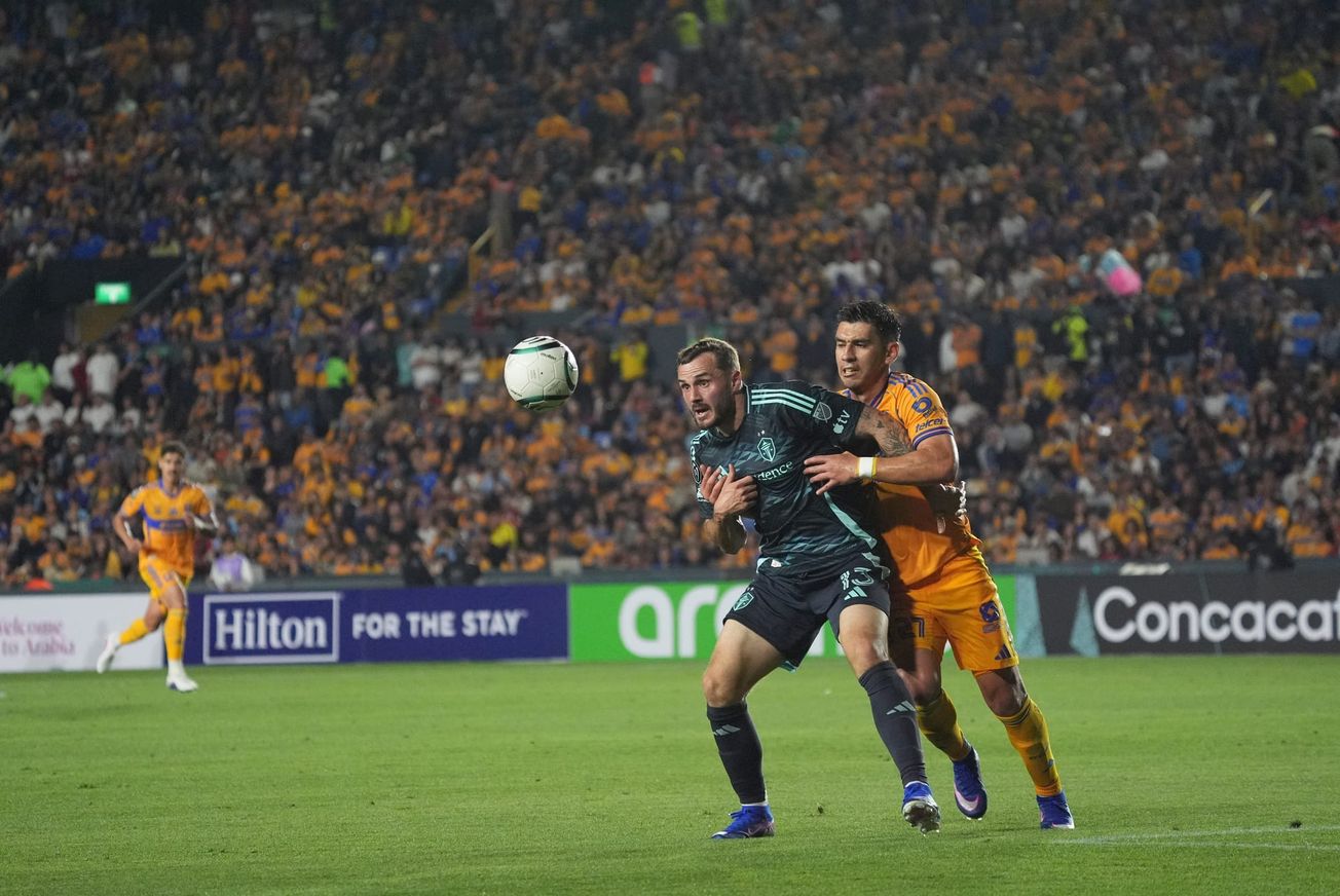 Jordan Morris wearing a navy blue Sounders kit holds off a Tigres defender with the ball in the air in front of him.