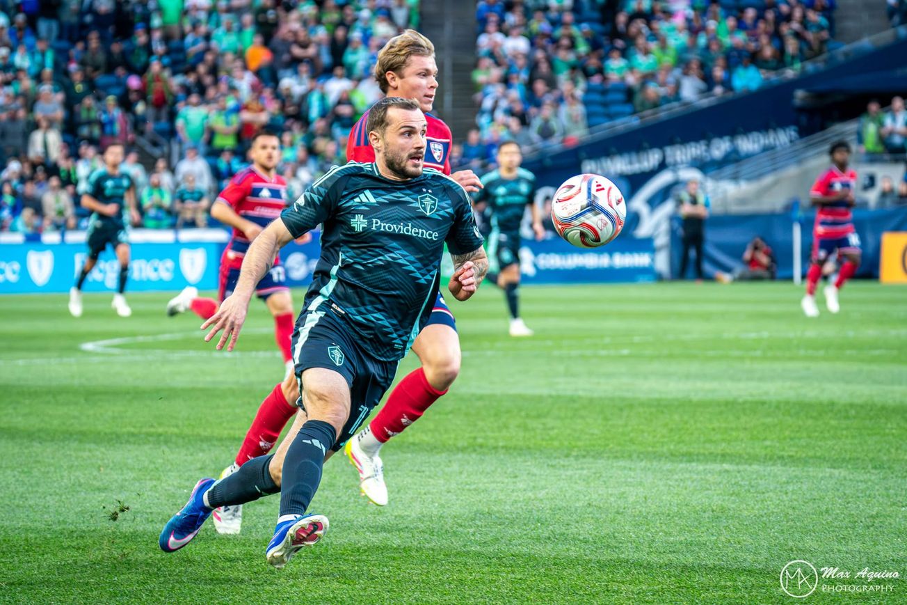 Jordan Morris in a dark blue Sounders kit runs with the ball in the air in front of hi