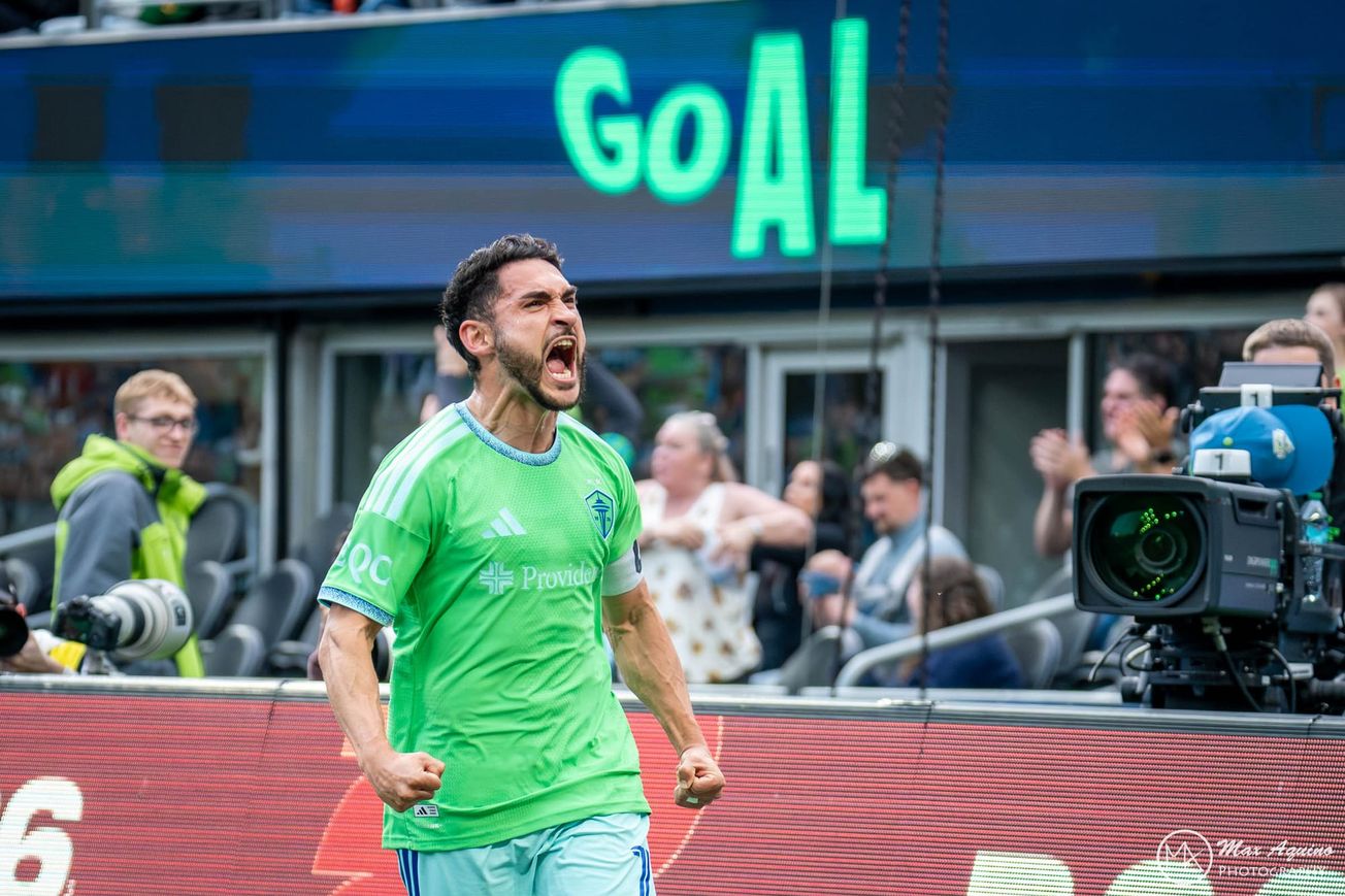 Cristian Roldan in a green Sounders jersey and light blue shorts celebrates a goal by flexing and yelling with a board reading “GOAL” behind him.