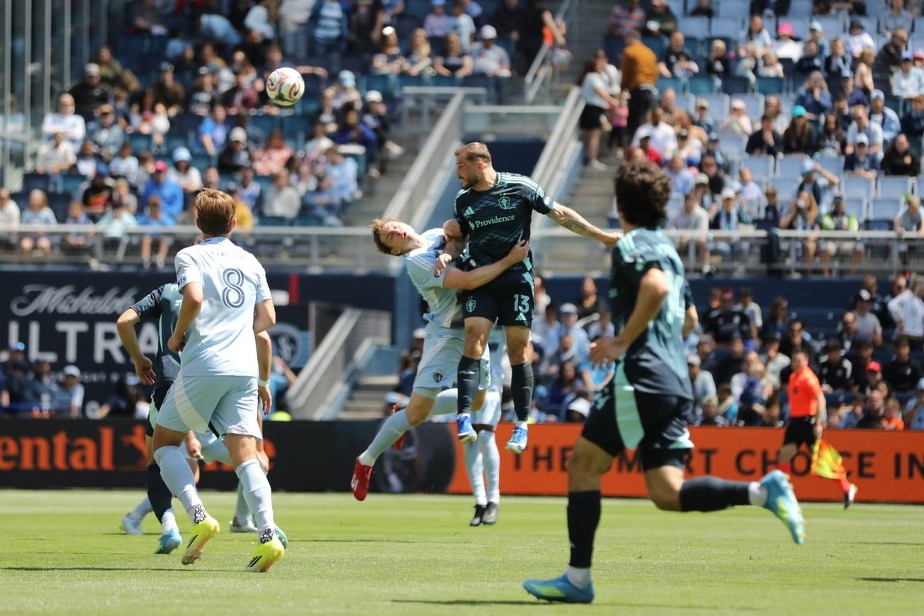Jordan Morris in a dark blue Sounders kit jumps to head the ball while a defender in light blue holds his waist