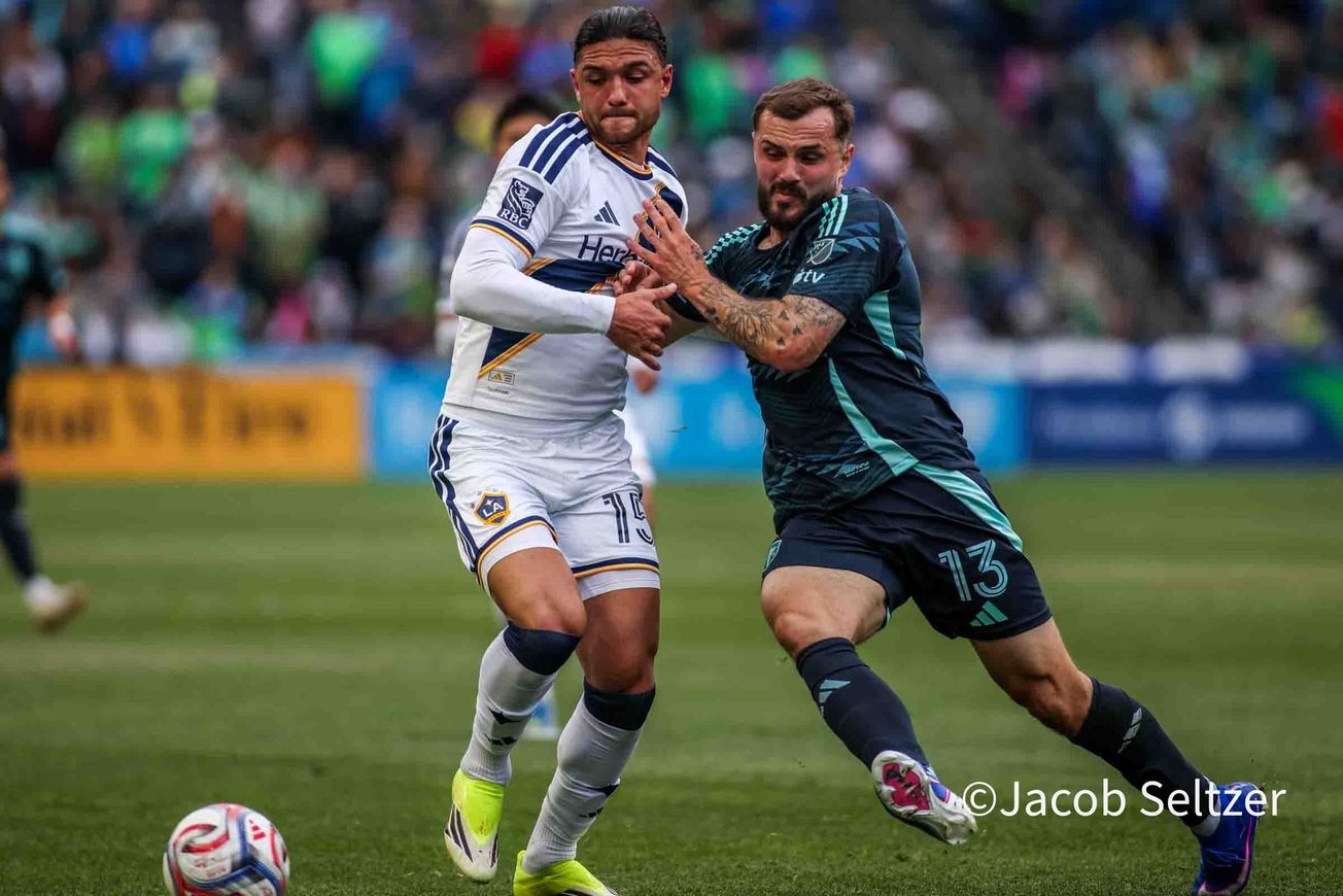 Jordan Morris in a dark blue Sounders kit pushes against a defender in a white LA Galaxy kit.