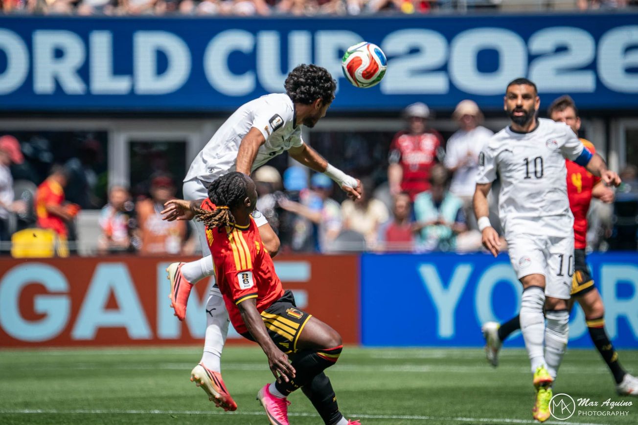 Jeremy Doku in red and black battles with an Egyptian player in white as the Egyptian player heads the ball with Mohamed Salah in the background wearing all white