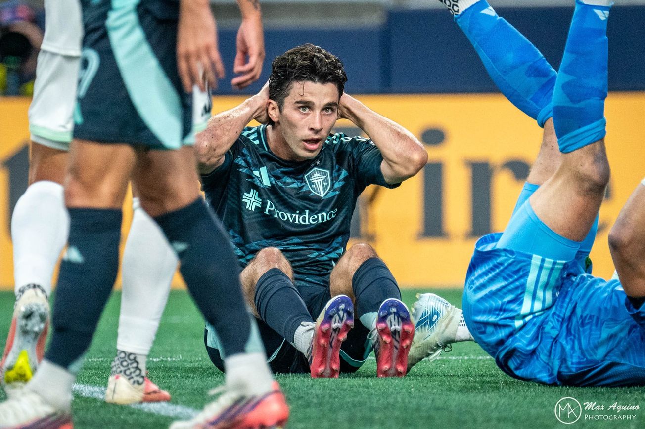 Paul Rothrock wears a dark blue Sounders kit while he sits on the ground with his head in his hands.