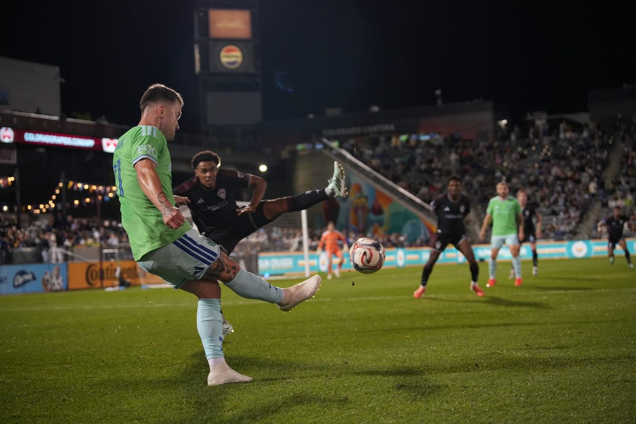 Albert Rusnák in a green Sounders jersey with light blue shorts sends in a cross defended by a Rapids player.