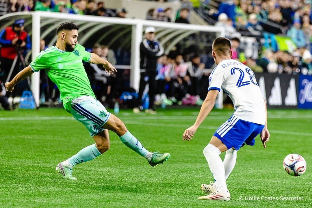 Alex Roldan in a green Sounders jersey and light blue shorts and socks kicks a ball ahead of a Montreal player in a white shirt and blue shorts.