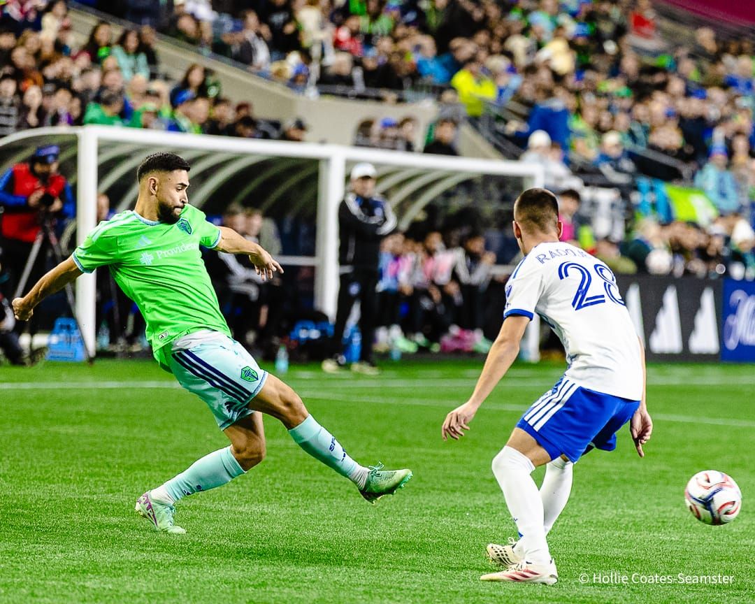 Alex Roldan in a green Sounders jersey and light blue shorts and socks kicks a ball ahead of a Montreal player in a white shirt and blue shorts.