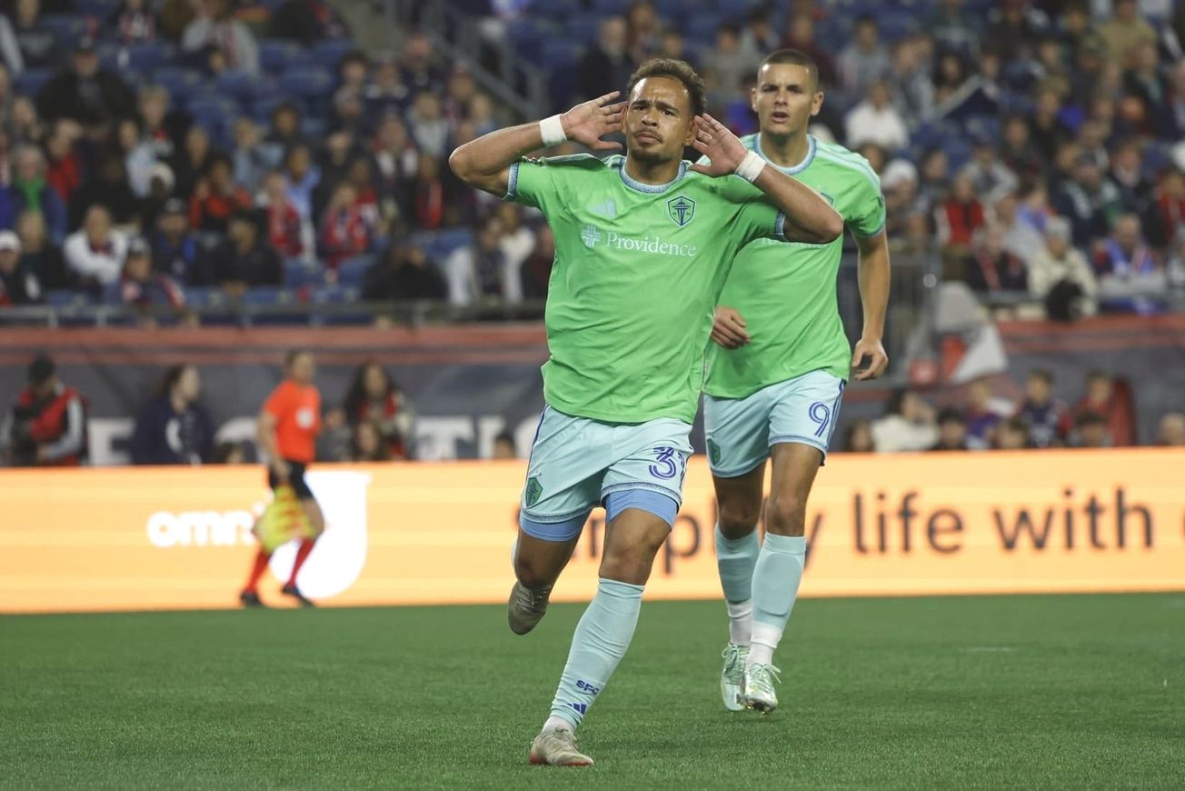 Hassani Dotson wearing a green Sounders jersey and light blue shorts and socks celebrates a goal with his hands to his ears