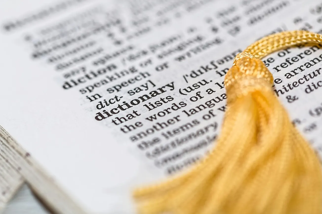 Close-up of a dictionary page focusing on the word "dictionary" with pronunciation and definition, accompanied by a yellow bookmark tassel.