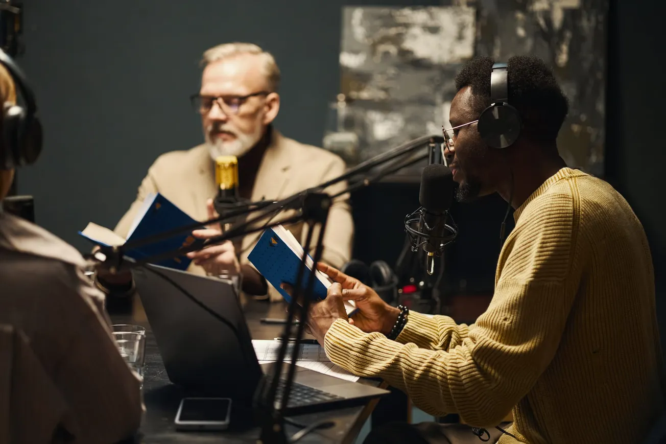 Podcast hosts recording in studio with microphones and headphones – Engaging in discussion while referencing notes and books.