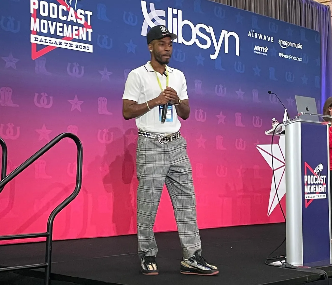 A person in a white shirt and plaid pants speaks on stage at Podcast Movement 2025 in Dallas. The backdrop features logos and bright pink and blue colors.
