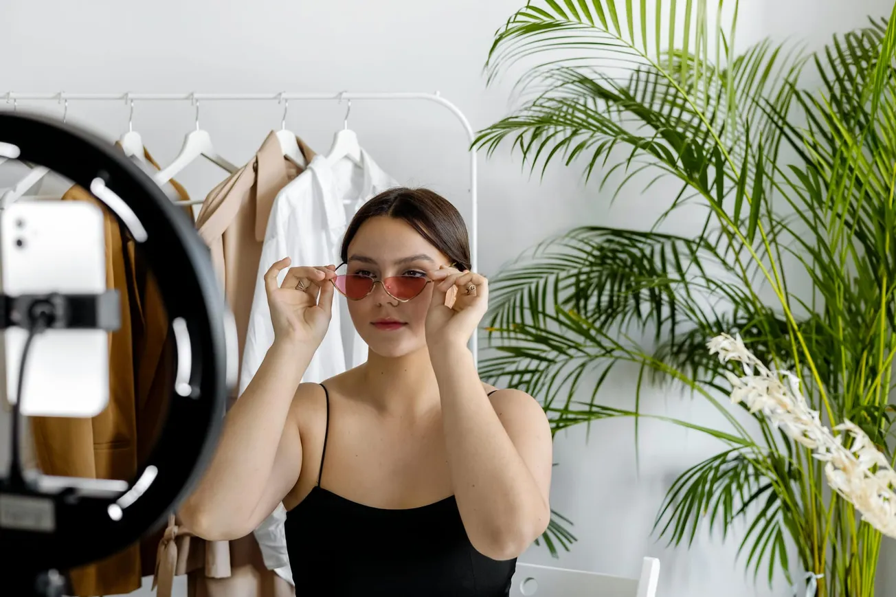 A woman in a black tank top adjusts red sunglasses, posing confidently in front of a smartphone on a ring light. Background includes clothes on a rack and a potted plant.