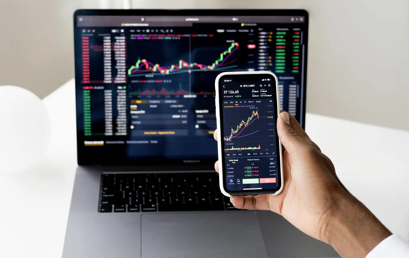 A person holds a smartphone displaying stock market charts in front of a laptop with similar charts.