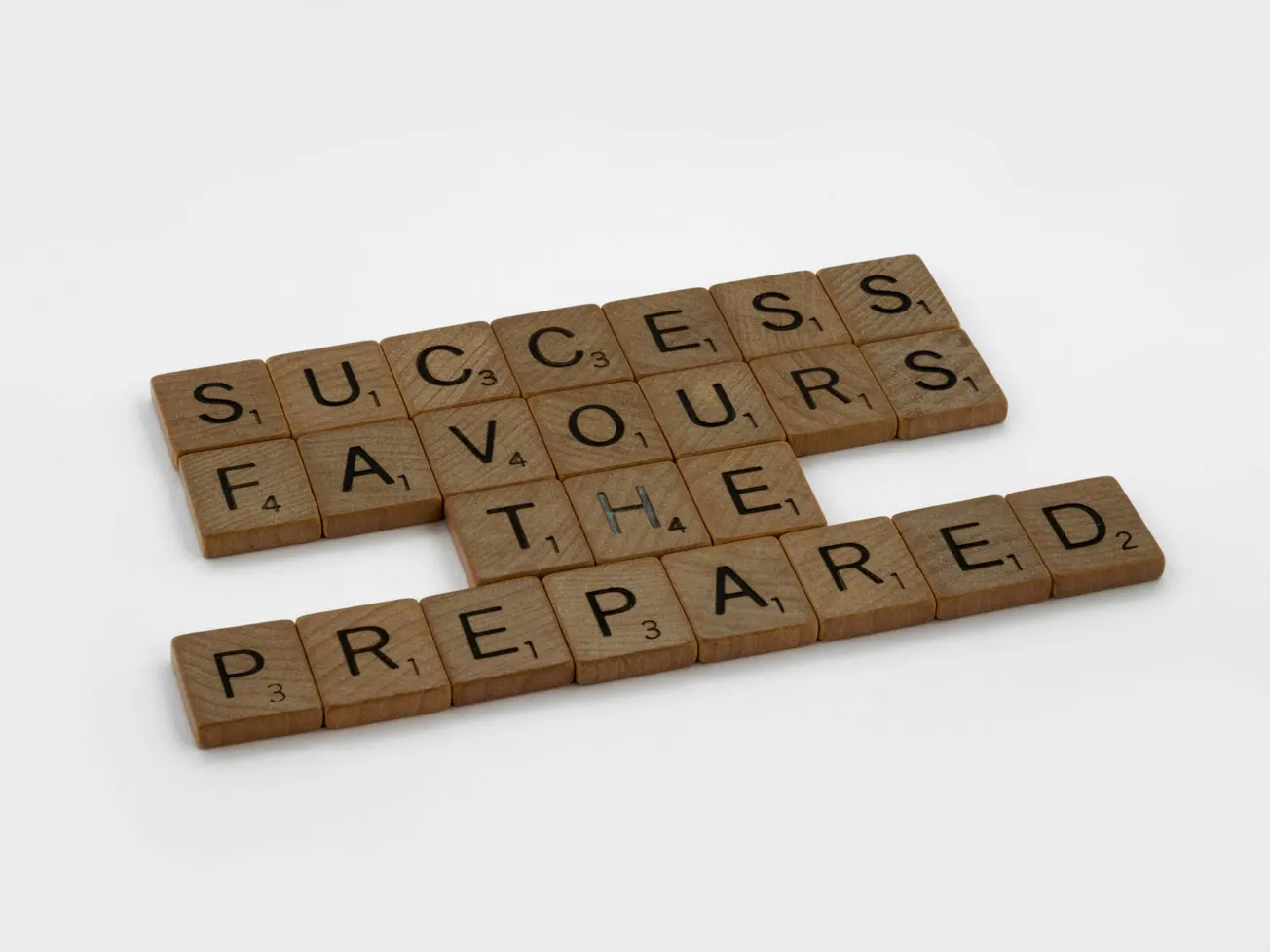 Wooden Scrabble tiles spell "Success favours the prepared." The tiles are neatly aligned on a gray background.