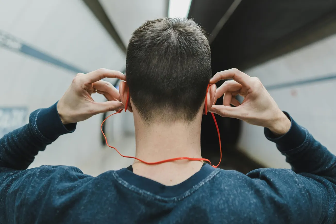 A person stands in a subway tunnel facing away, wearing a navy sweater, with red earphones being adjusted. 